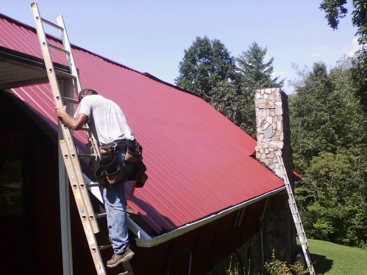 Roofer on a ladder beside a red metal roof and stone chimney — Soffit & Fascia Repair by Roofing Brothers in Beaver Springs